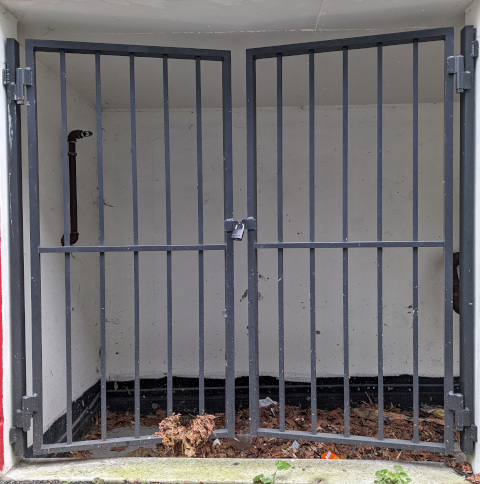 A pair of iron gates, padlocked together, guard some kind of lil alcove full of dead leaves and garbage